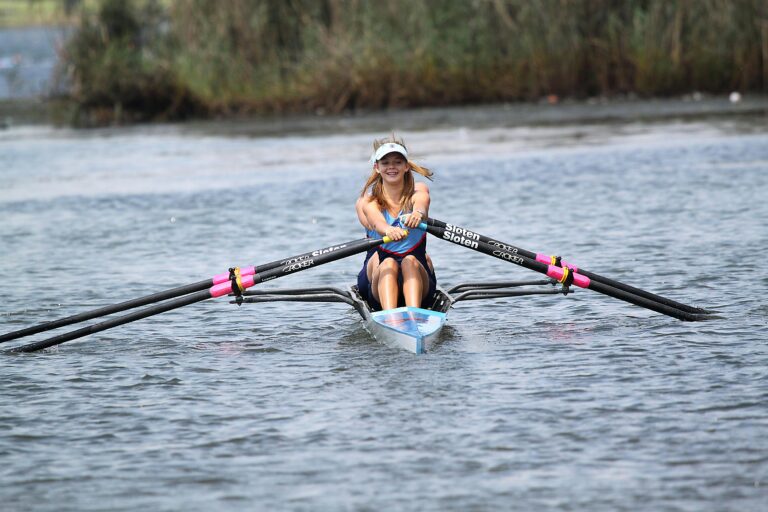 Smiling woman rowing a scull on a calm lake, enjoying outdoor water sports.